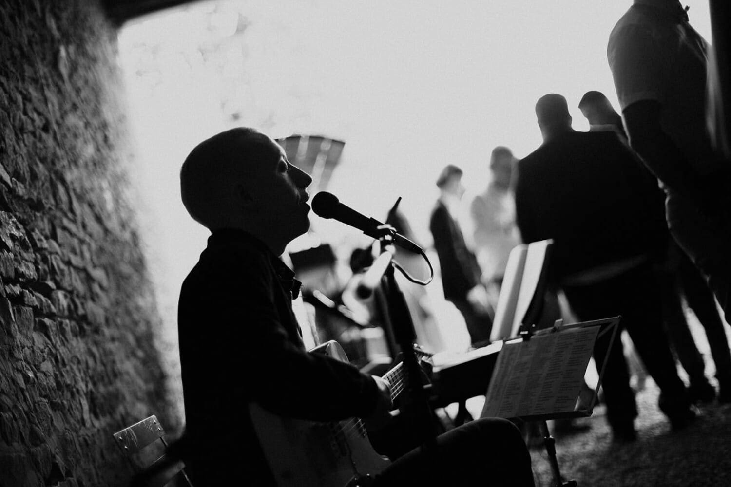 Chanteur et guitariste en noir et blanc pendant un cocktail de mariage, silhouettes d’invités floutées en arrière-plan.