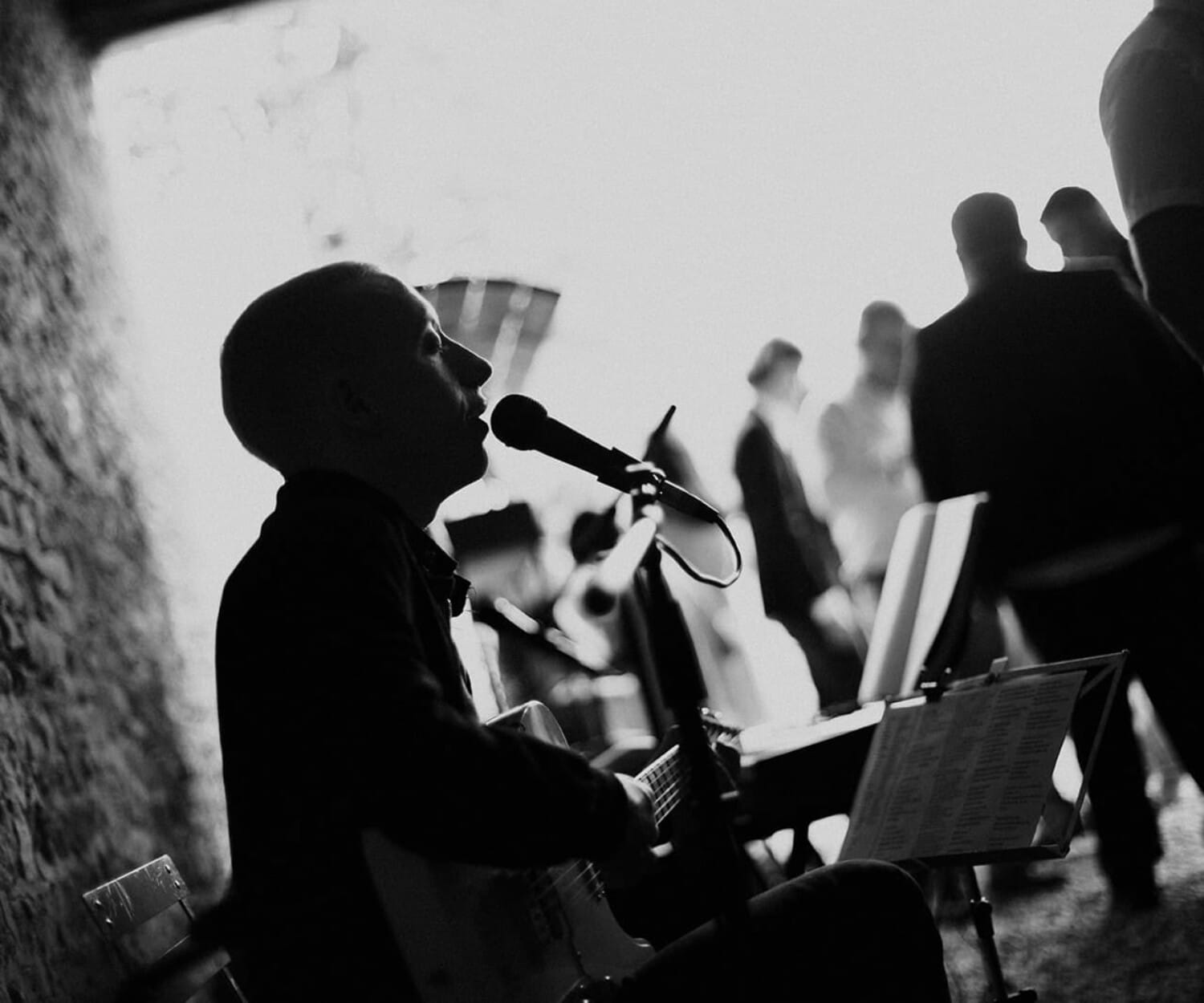 Chanteur et guitariste en noir et blanc pendant un cocktail de mariage, silhouettes d’invités floutées en arrière-plan.