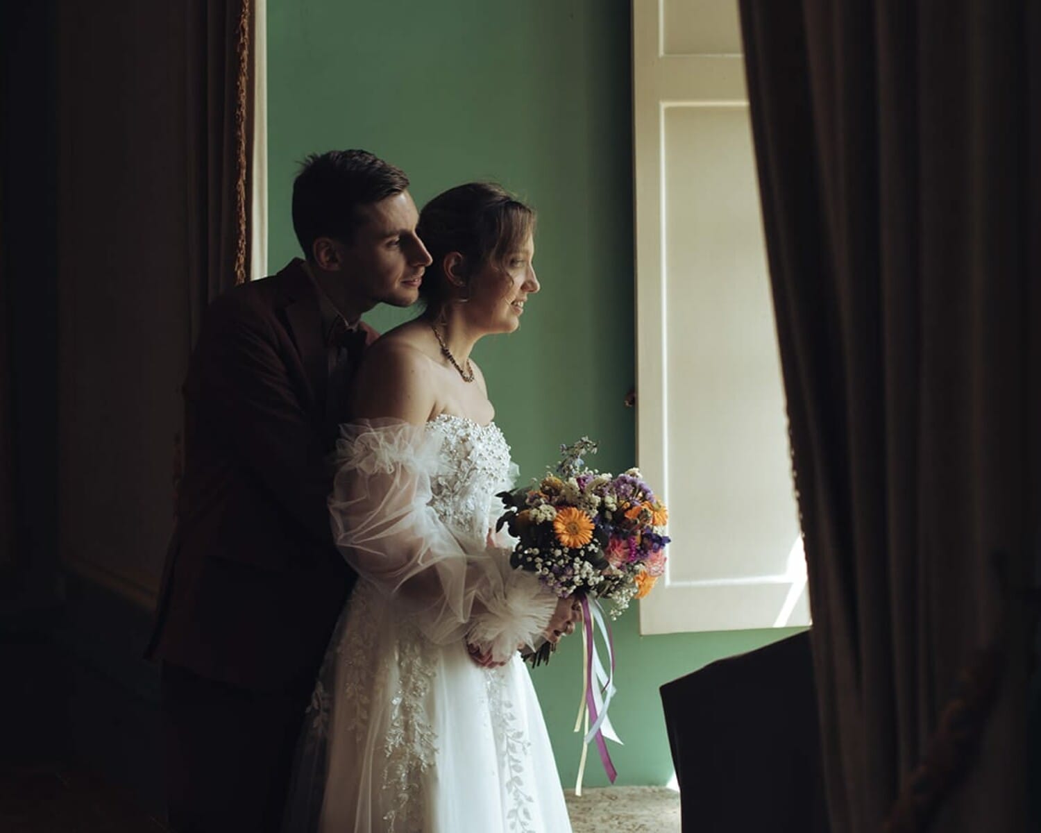 Couple de jeunes mariés regardant par la fenêtre, bouquet en main, baigné·es par la lumière douce d’Aix-les-Bains.