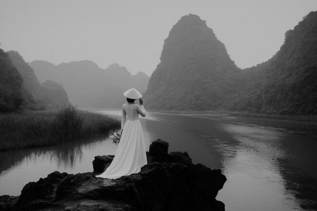 Vietnamese bride in white áo dài and conical hat standing on a rock by a river, surrounded by dramatic limestone mountains in black and white