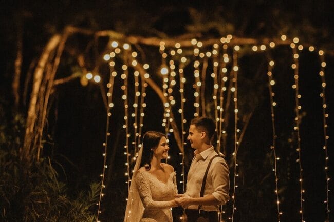 Outdoor night wedding scene with bride and groom under string lights, surrounded by candles and a cake table in Vietnam.