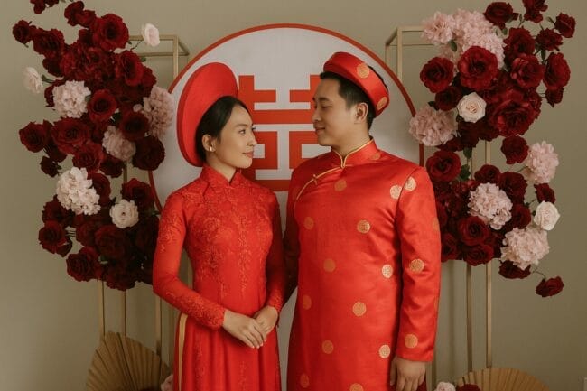 Vietnamese bride and groom in traditional red áo dài during their wedding celebration, standing in front of a floral backdrop and double happiness symbol.