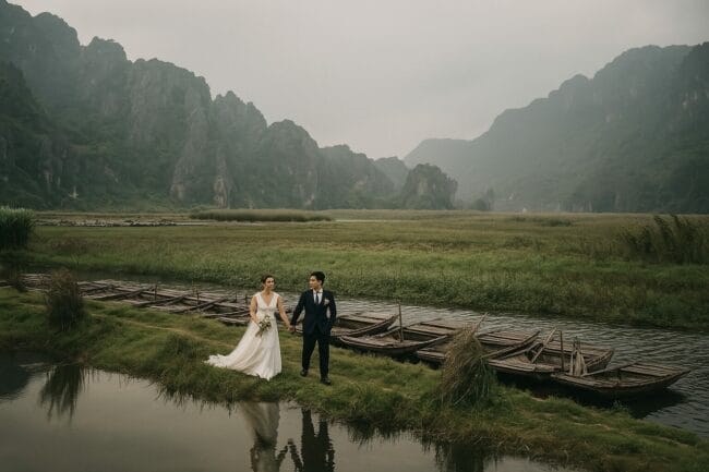 Vietnamese bride and groom walking hand in hand near traditional wooden boats in Ninh Binh, with dramatic limestone mountains in the background