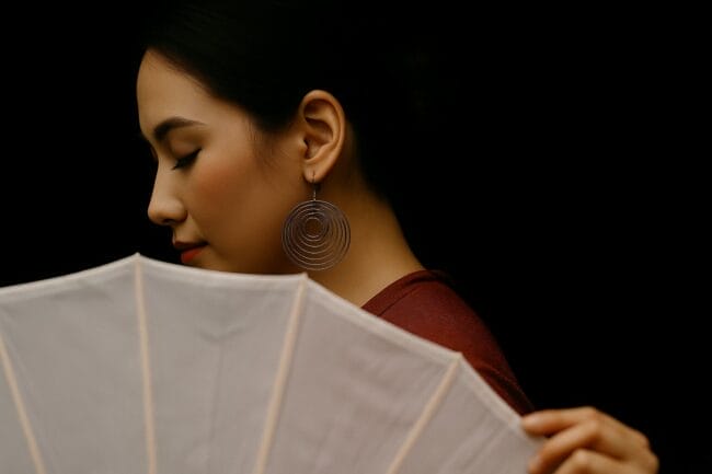 Vietnamese woman in red dress holding a white fan, with spiral earrings and soft lighting – bridal or portrait session
