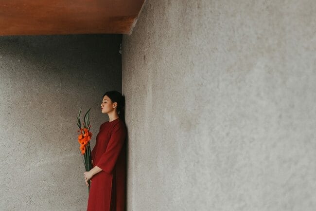 Vietnamese woman in red áo dài holding orange flowers, standing against a minimalist concrete wall in natural light