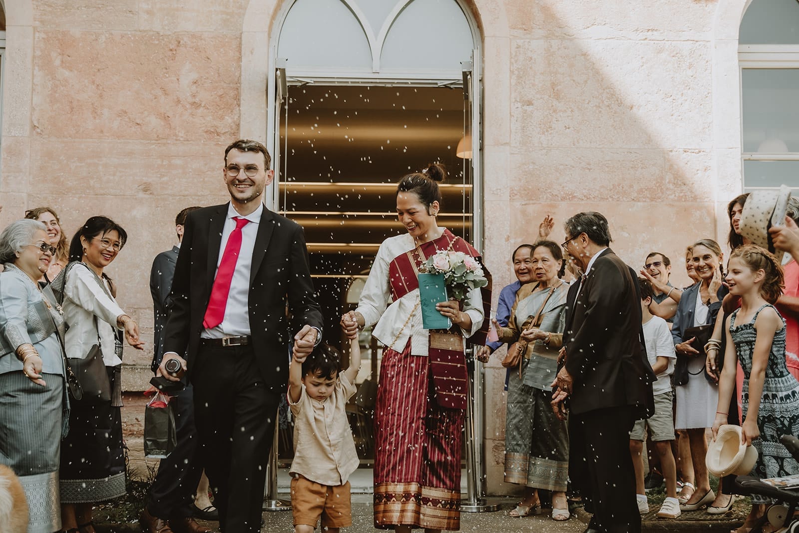 Couple mixte sortant de la mairie de Caluire-et-Cuire avec leur enfant, entourés de leur famille, la mariée portant une tenue traditionnelle du Laos