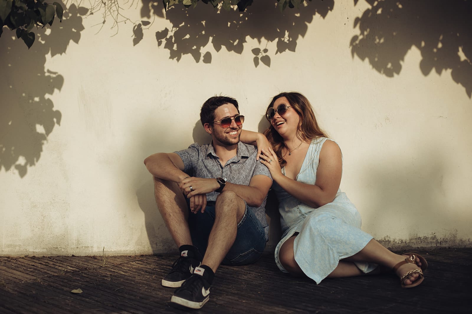 Couple assis contre un mur à l’ombre des feuilles, riant ensemble lors d’une love session à Lyon