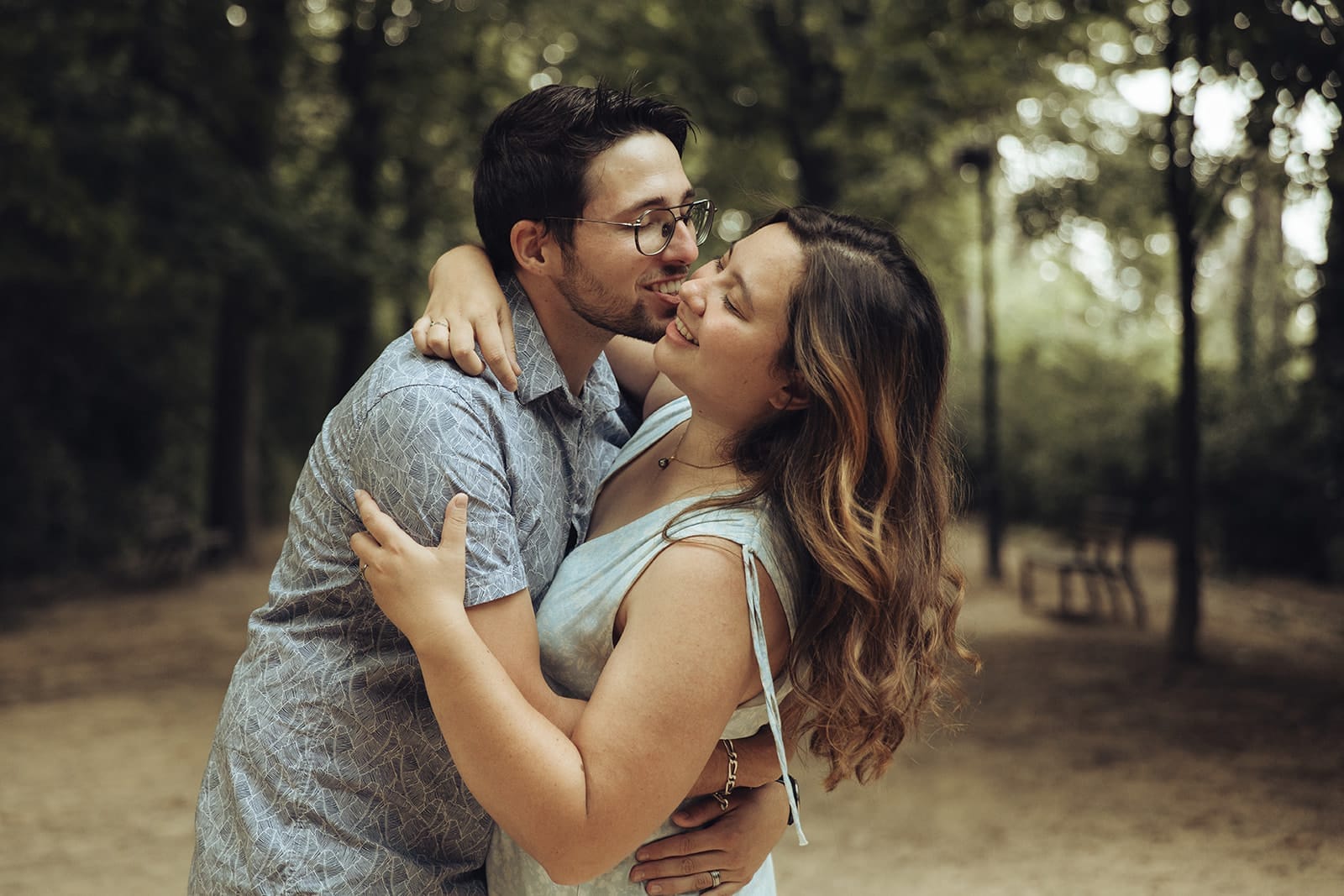 Couple se serrant tendrement dans une allée arborée, sourire et baiser sur le front
