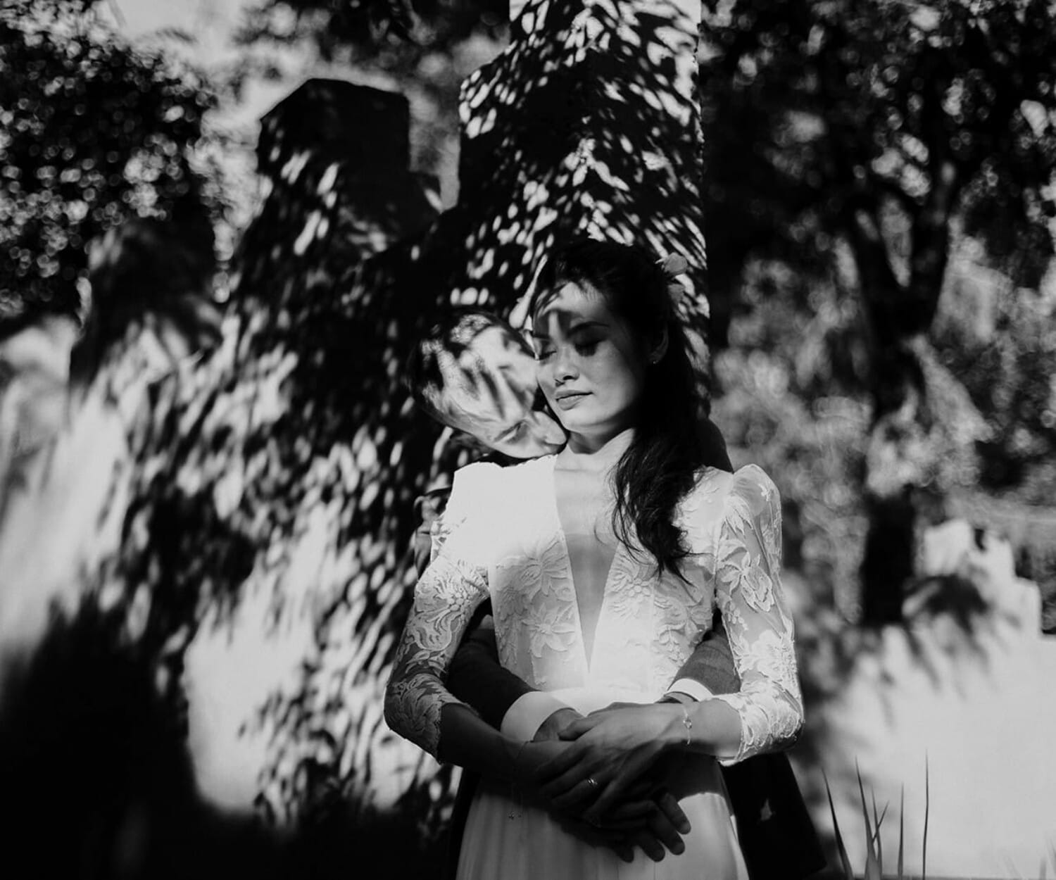 Mariée en robe à manches longues, debout dans les bras de son compagnon, baignée d’ombres et de lumière sous les arbres.