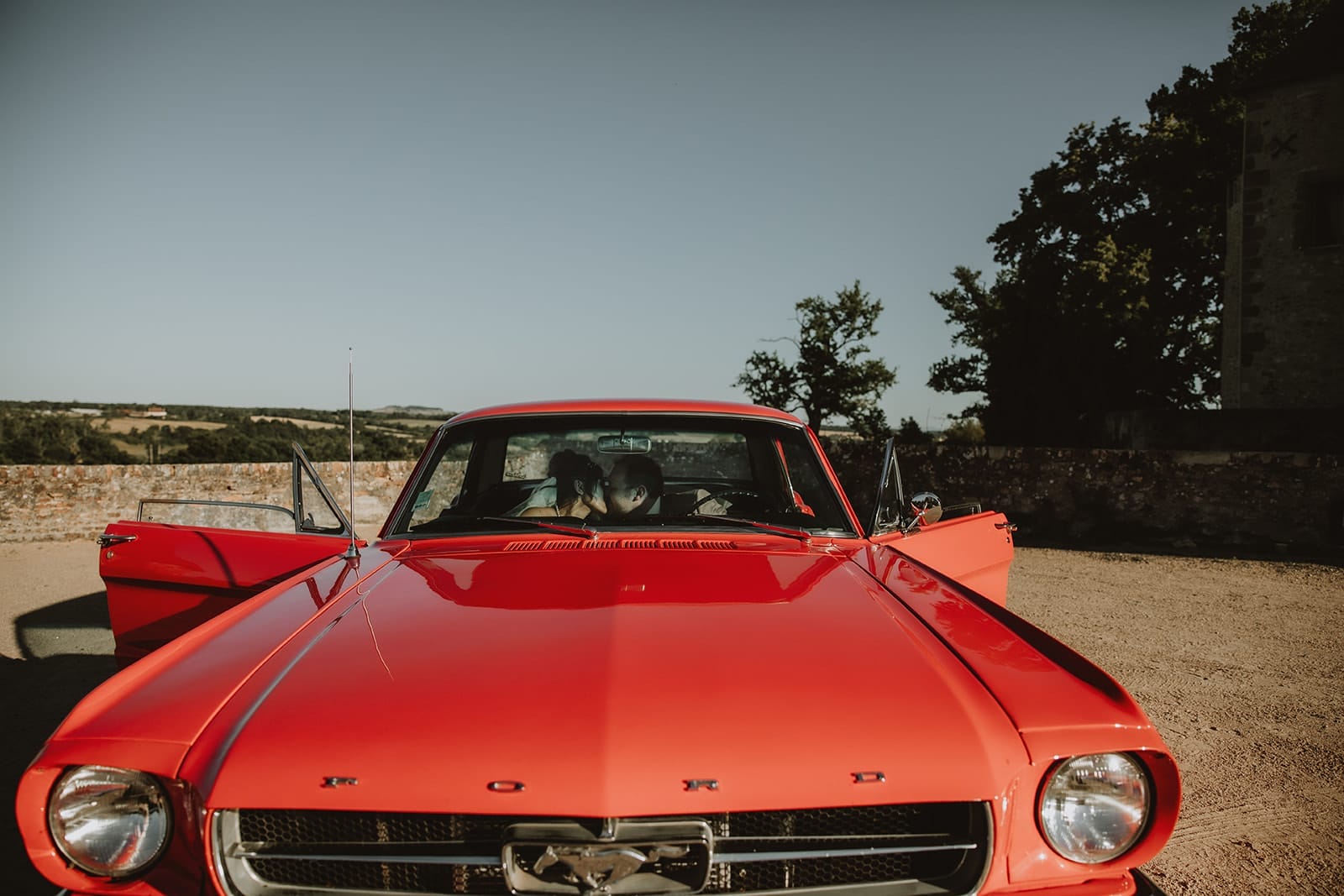 Mariés assis dans une Mustang rouge, portes ouvertes, devant le Château de Beauvoir