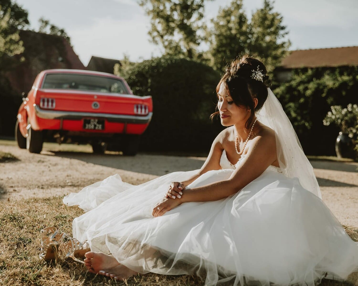 Mariée assise pieds nus devant une Mustang rouge, robe étalée au sol