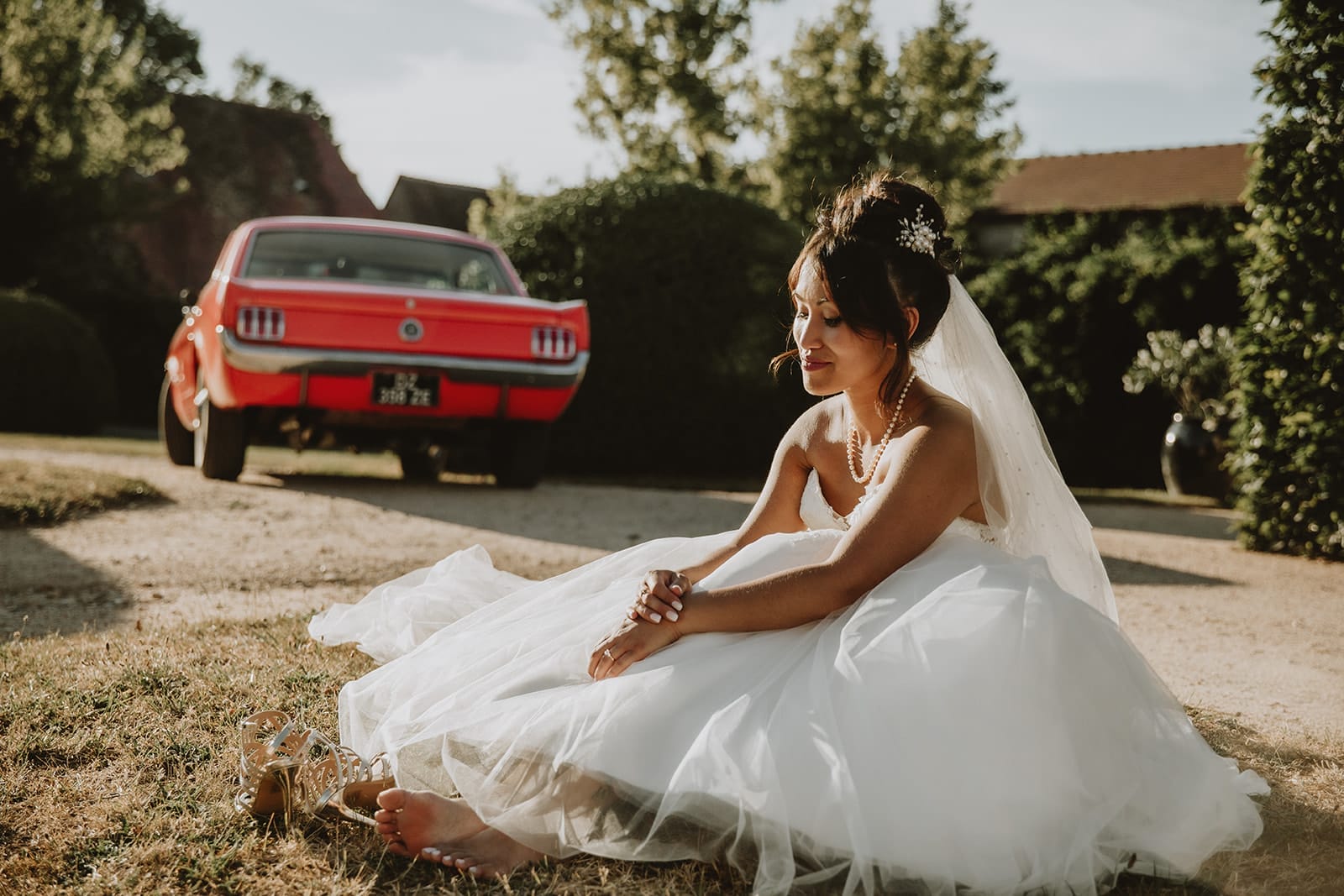 Mariée assise pieds nus devant une Mustang rouge, robe étalée au sol